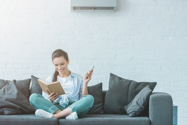 woman reading book while turning on air conditioner with remote control - Photo, image