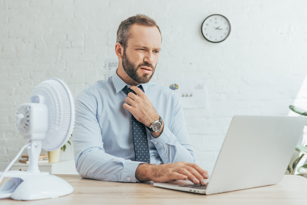 homem de negócios barbudo trabalhando com laptop no espaço de trabalho com ventilador elétrico
  - Foto, Imagem