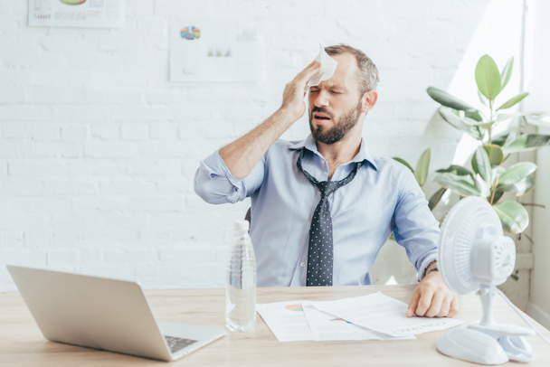 hombre de negocios cansado con dolor de cabeza sentado en el lugar de trabajo con ventilador eléctrico, botella de agua, papeleo y portátil
 - Foto, Imagen