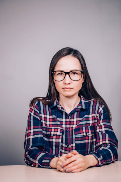 Serious business woman with glasses sitting at a table in a shirt on a gray background - Фото, изображение