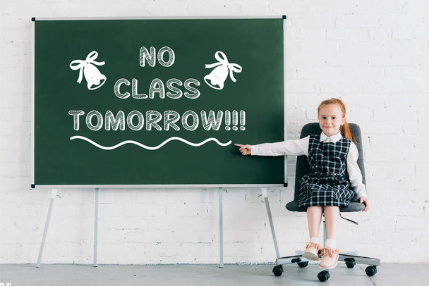 adorável colegial sorrindo para a câmera e apontando para quadro-negro com letras "sem aula amanhã"
 - Foto, Imagem