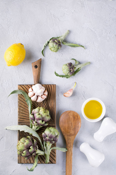 Alcachofas verdes maduras frescas en tabla de cortar lista para cocinar e ingredientes ajo, limón y aceite de oliva
 - Foto, Imagen