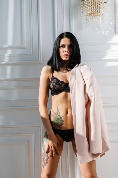 Fashion studio shot of beauty brunette woman with tattoo on body. Beautiful female model in stylish black lingerie standing in luxury apartments interior - Fotó, kép
