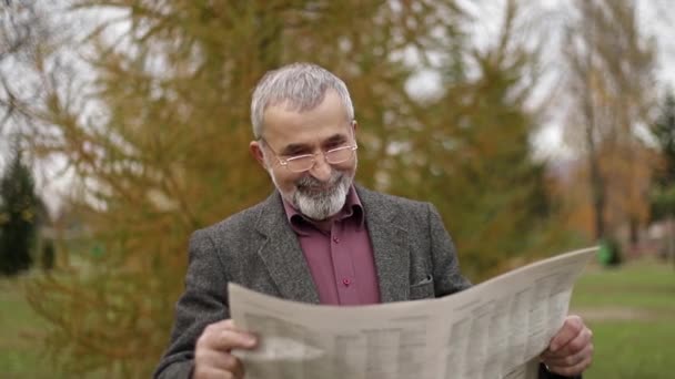 Miły dziadek z pięknej brody w szarą kurtkę w parku i czyta gazetę - Materiał filmowy, wideo