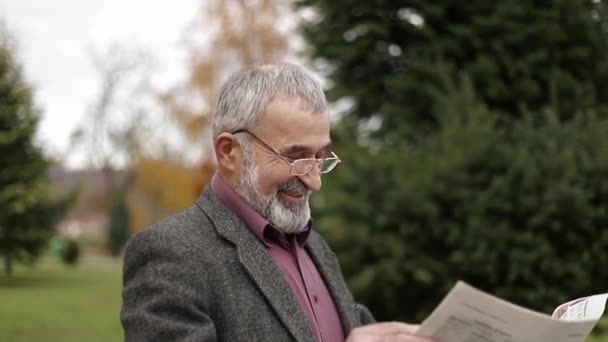A nice grandfather with a beautiful beard in a gray jacket in the park and reads a newspaper - Imágenes, Vídeo