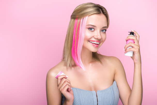 χαμογελώντας νεαρή γυναίκα με πολύχρωμα σκέλη της τρίχας και σπρέι βαφής που βλέπουν τα φωτογραφικών μηχανών που απομονώνονται σε ροζ - Φωτογραφία, εικόνα