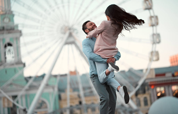 Love is in the air! Cute romantic couple spending time together in the city. Handsome bearded man and attractive young woman are in love. Hugging, kissing and having fun on the Ferris wheel background. - Fotografie, Obrázek