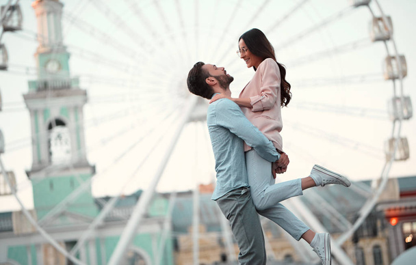 Love is in the air! Cute romantic couple spending time together in the city. Handsome bearded man and attractive young woman are in love. Hugging, kissing and having fun on the Ferris wheel background. - Foto, imagen