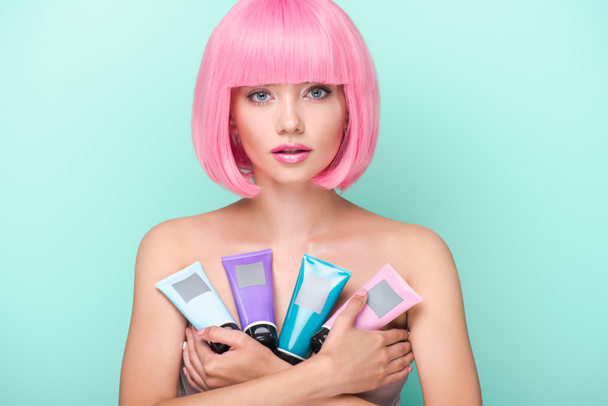 beautiful young woman with pink bob cut holding various tubes of coloring hair tonics looking at camera isolated on turquoise - Photo, Image