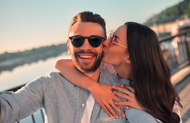 Love is in the air! Cute romantic couple spending time together in the city and making selfie. Handsome bearded man and attractive young woman are in love. Hugging, kissing and having fun during the sunset. - Фото, изображение