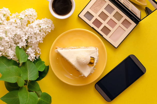 Sponge cake with hazelnut, cheese-coconut cream on natural cream and marmalade layer of passion fruit, cup of cocoa drink, mobile phone and lilac tree branch flowers isolated on yellow background. - Foto, Imagen