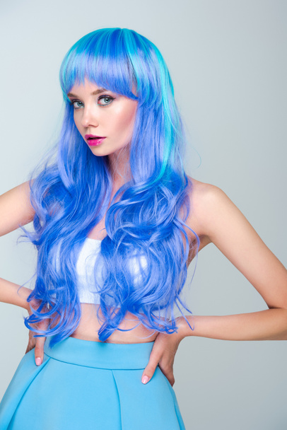 serious young woman with bright blue hair standing with arms akimbo and looking at camera isolated on grey - Foto, afbeelding