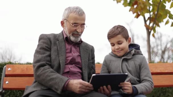 Grandpa and his grandson spend time together in the park. They are sitting on the bench and use the tablet - Footage, Video