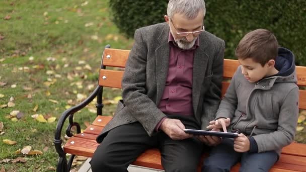 おじいちゃんと孫、一緒に公園で時間を過ごします。彼らはベンチに座っているし、タブレットを使用 - 映像、動画
