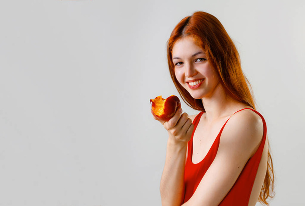 Sexy slim woman in red swimsuit posing and eating peach in studio. Full length fashion portrait of beautiful girl with long wavy red hair. Copy space, gray background - Photo, Image