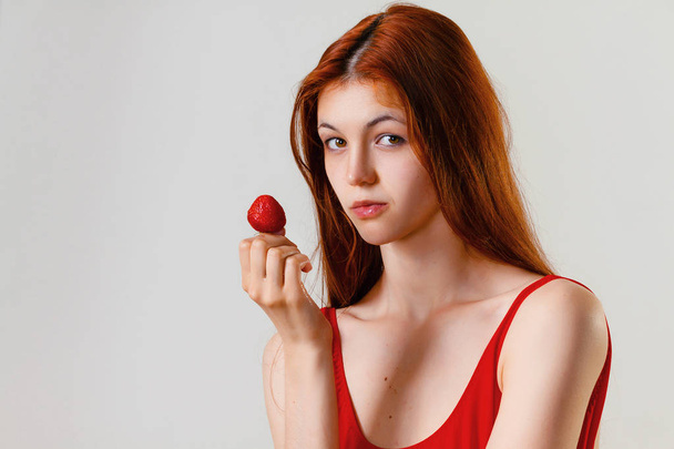 Sexy femme mince en maillot de bain rouge posant et tenir la fraise en studio. Portrait de mode pleine longueur de belle fille aux longs cheveux roux ondulés. Espace de copie, fond gris
 - Photo, image