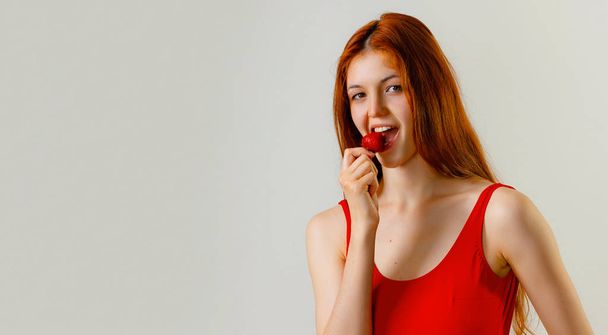 Sexy mujer delgada en traje de baño rojo posando y comiendo fresa en el estudio. Retrato de moda de longitud completa de hermosa chica con el pelo largo y ondulado rojo. Copiar espacio, fondo gris
 - Foto, imagen