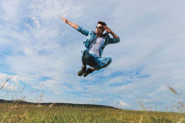 young man with sunglasses salutes with his hands to side whle jumping outside on a grass field - Foto, Bild