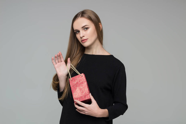 Genç model, uzun saçlı, siyah elbiseli sarışın kız elinde kırmızı kağıt hediye paketi tutuyor. - Fotoğraf, Görsel