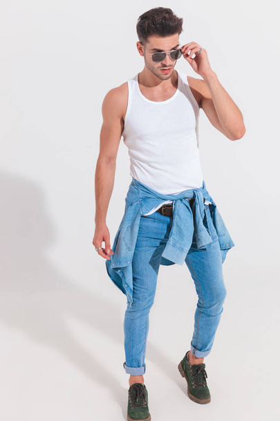 attractive man with wearing undershirt and denim shirt around waist fixes sunglasses and looks to side while standing on white background in projector light, full length picture - Foto, Imagem