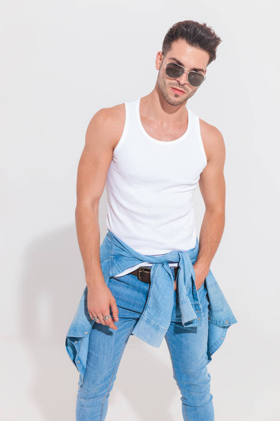 портрет соблазнительного случайного мужчины в джинсовой рубашке вокруг талии, стоящего с рукой в кармане в центре внимания возле белой стены. На нем пара солнцезащитных очков и джинсы
 - Фото, изображение