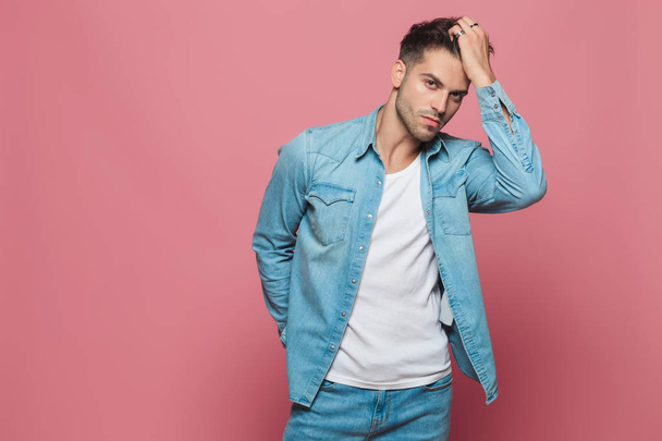 portrait of casual man wearing denim shirt fixing his hair while standing on pink background - 写真・画像