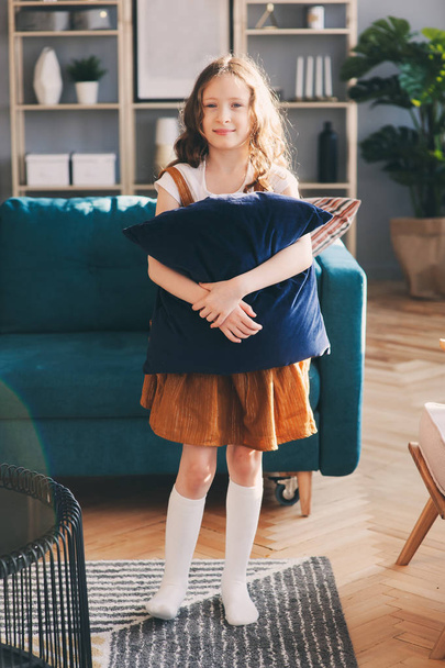 happy cheerful kid girl posing with pillow, cozy couch behind in modern living room. Buying new home, moving to big apartments, house rental concept. - 写真・画像