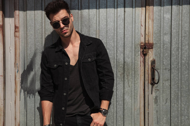 handsome man wearing black clothes and sunglasses standing near garage door with hand in pocket on a sunny day, portrait picture - 写真・画像