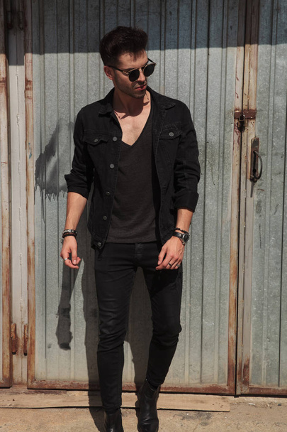 man wearing black shirt and sunglasses is standing in front of metal wall and looking down to side, full length picture - Zdjęcie, obraz