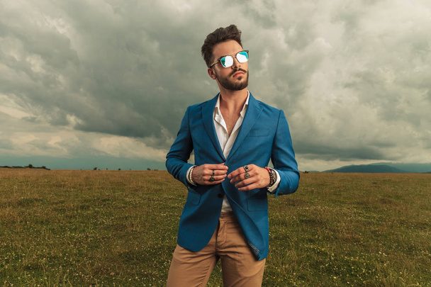 hombre casual inteligente con gafas de sol abotonando su traje azul en el tiempo nublado fuera en un campo, retrato de la imagen
 - Foto, imagen