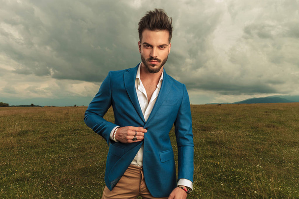 smart casual man buttoning his blue suit on cloudy weather while standing on a field, portrait picture - Photo, Image