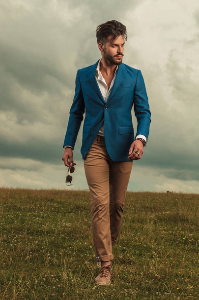 smart casual man walking and looking to side while holding sunglasses on grass field on a cloudy weather, full body picture - Foto, Bild