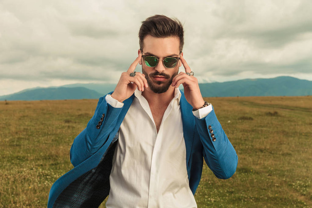 portrait of smart casual man standing outdoor in a grass field with clouds in the sky and fixing sunglasses - Photo, Image