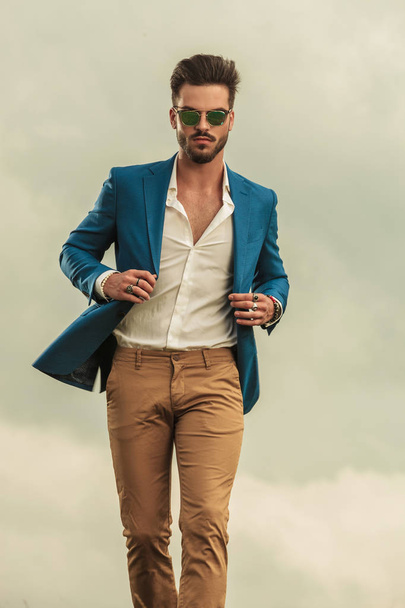 сексуальна людина тримає синій костюм коміром і носить сонцезахисні окуляри, що ходять на хмарах фоні
,  - Фото, зображення