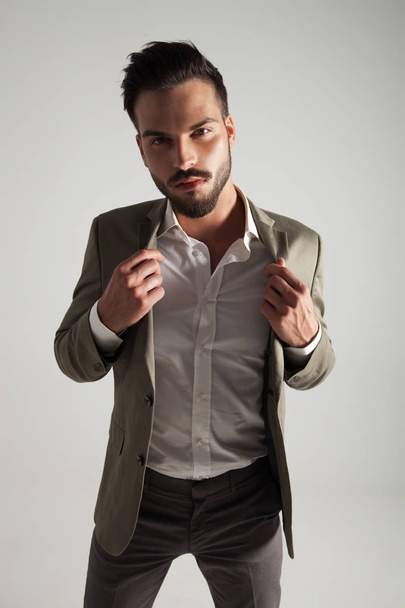 seductive elegant man holds his green suit's collar while standing on light grey background, portrait picture - Foto, Imagem
