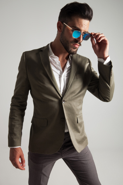 κομψό άνδρα σε πράσινο κοστούμι διορθώνει τα γυαλιά ηλίου και μοιάζει πλευρά ενώ στέκεται στο ανοιχτό γκρι φόντο, φωτογραφία πορτραίτου - Φωτογραφία, εικόνα
