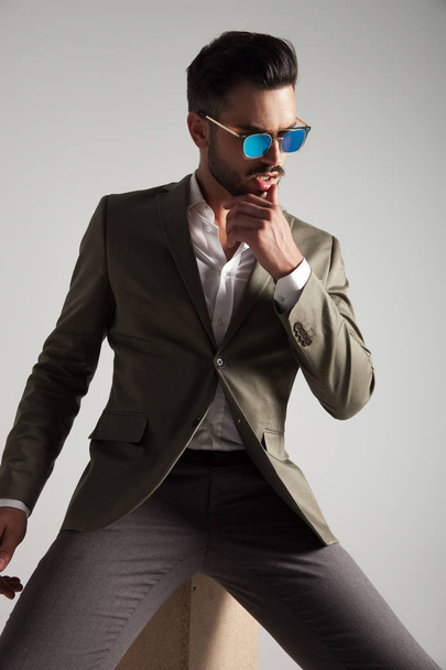 pensive man wearing sunglasses and green suit sitting on a wooden crate and looking to side, portrait picture - Foto, afbeelding
