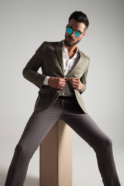 handsome man with sunglasses buttoning his green suit and looking down to side while sitting on a wooden box on light grey background - Фото, изображение