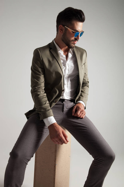 elegant man wearing green suit and sunglasses looks down to side while sitting on a wooden box on light grey background - Foto, imagen