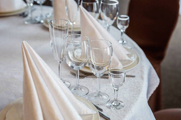 Glasses, napkin folded in a pyramid, served for dinner in restaurant with cozy interior. Wedding decorations and items for food, arranged by the catering service on a large table covered with white tablecloth - Photo, Image