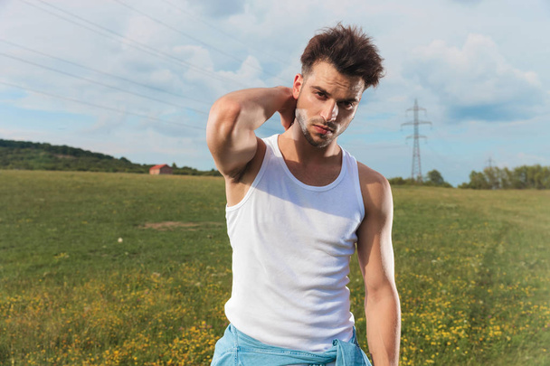 young man in undershirt arranges his hair while standing outside in a grass field - Photo, image