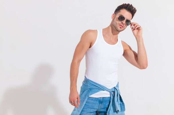 portrait of young man in undershirt fixing his sunglasses while standing on white background and leaning to side - Foto, Imagem