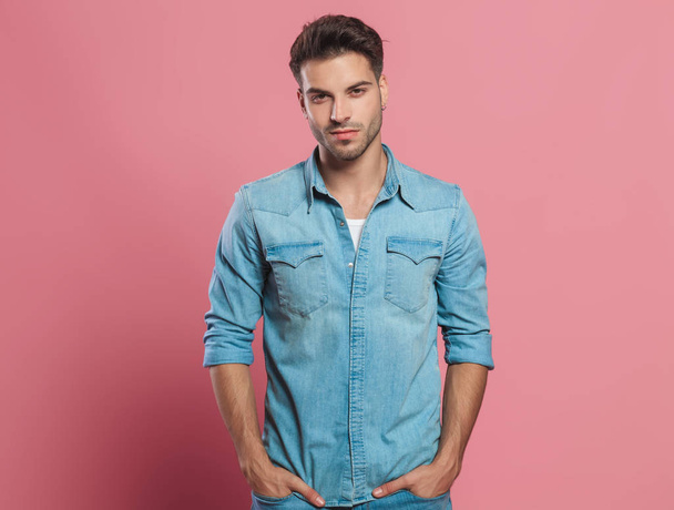portrait of relaxed young man wearing a denim shirt standing on red background with hands in pockets - Foto, Imagem