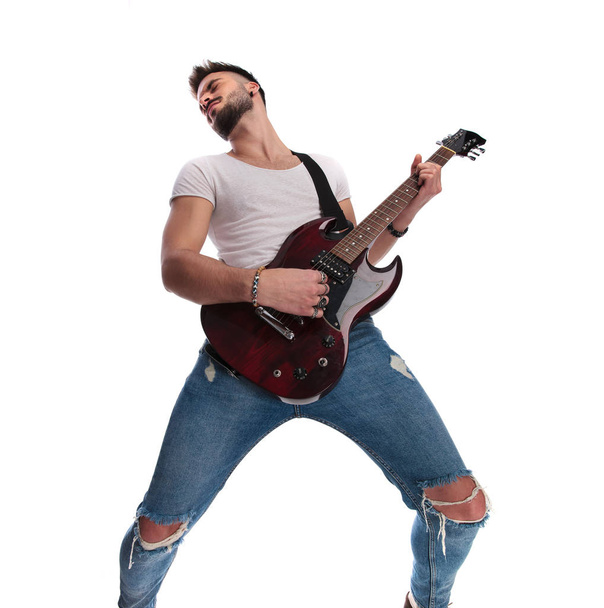 emocionado joven guitarrista tocando la guitarra y recostado hacia atrás, mientras que de pie sobre fondo blanco
 - Foto, Imagen