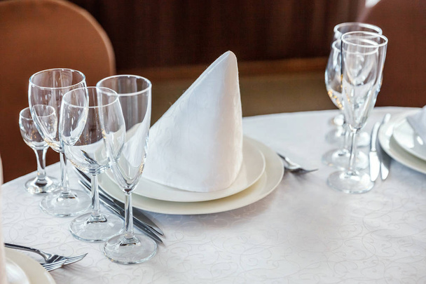 Okulary, serwetka składany w piramidzie, serwowane w porze kolacji w restauracji z przytulnym wnętrzem. Ślubne ozdoby i przedmioty do żywności, ułożone przez usługi cateringowe na duży stół przykryty białym obrusem - Zdjęcie, obraz