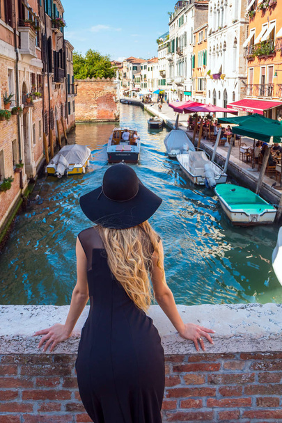Venice, Italy - September 25, 2016: Elegant woman with black sunhat enjoys the view to a canal with passing by gondola in Venice, Italy - Photo, Image