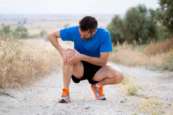 young sport man with strong athletic legs holding knee with his hands in pain after suffering muscle injury during a running workout training in asphalt road in muscular or ligament injury concept. - Valokuva, kuva