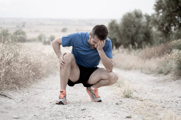young sport man with strong athletic legs holding knee with his hands in pain after suffering muscle injury during a running workout training in asphalt road in muscular or ligament injury concept. - 写真・画像