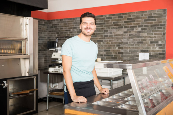 Jeune serveur dans un magasin de pizza debout derrière un comptoir souriant regardant la caméra
 - Photo, image