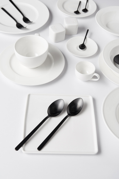 selective focus of different plates, cup, bowl, salt pot and pepper caster, forks with spoons on white table - Fotografie, Obrázek